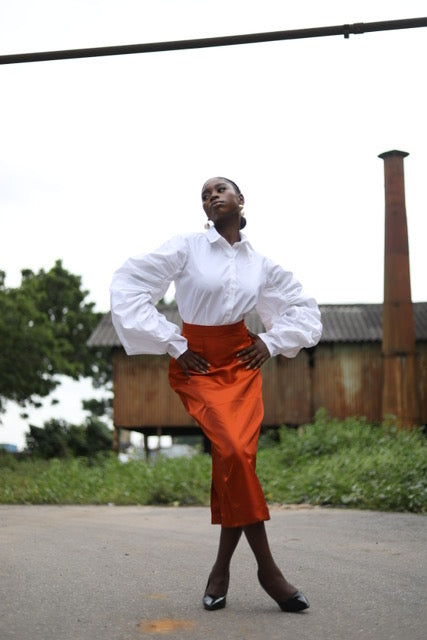 A model striking a pose in an outdoor setting, wearing a sophisticated white blouse with voluminous sleeves paired with a high-waisted orange midi skirt. The outfit is complemented by black heels. The backdrop includes rustic buildings and greenery, creating a contrast with the elegant fashion ensemble. Ideal for showcasing modern, bold fashion styles.