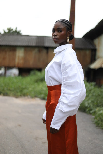 A model striking a pose in an outdoor setting, wearing a sophisticated white blouse with voluminous sleeves paired with a high-waisted orange midi skirt. The outfit is complemented by black heels. The backdrop includes rustic buildings and greenery, creating a contrast with the elegant fashion ensemble. 