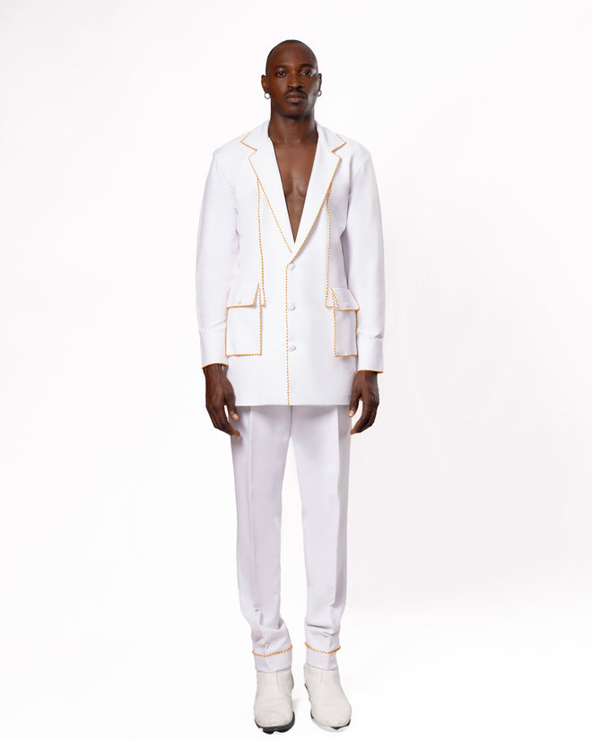 Man wearing a stylish white suit with gold stitching, standing against a plain background. The contemporary suit design features a deep V-neck blazer, complemented by matching tailored pants and white shoes. Ideal for high-fashion and modern style themes.